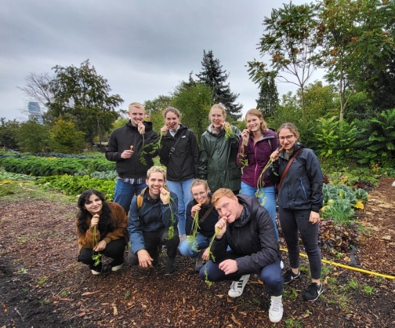 Eine Gruppe junger Menschen steht auf einem Feldweg vor einem Gemüsebeet. Sie tragen wetterfeste Kleidung und halten frisch geerntete Möhren in den Händen, einige beißen in das Gemüse. Im Hintergrund sind verschiedene grüne Pflanzen und Bäume zu sehen, der Himmel ist bewölkt.