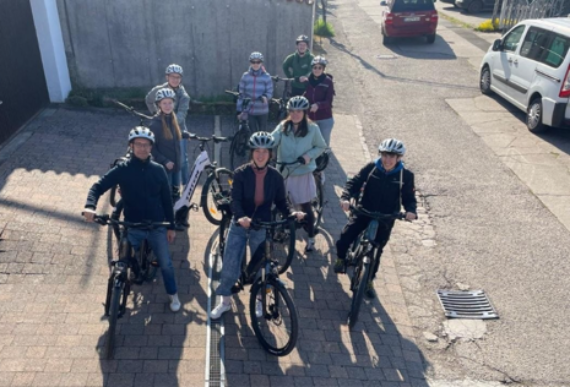 Mehrere Personen mit Fahrradhelmen stehen mit Fahrrädern auf einem gepflasterten Hof. Die Gruppe blickt nach oben in Richtung Kamera. Im Hintergrund sind ein grauer Zaun, ein geparkter weißer Transporter und ein rotes Auto auf einer asphaltierten Straße zu sehen. Die Szene wirkt sonnig, die Schatten sind klar erkennbar.